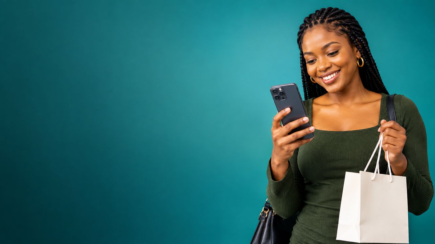 A smiling woman looking at her phone and holding a shopping bag against a teal background